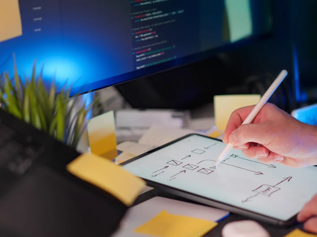 A hand sketching a user flow on a tablet beside a code monitor